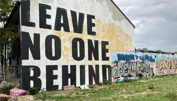 Eine Hausfassade, auf die der Schriftzug &quot;Leave No One Behind&quot; aufgemalt wurde. Rechts schlie?t sich eine Mauer mit bunten Graffitis an. 